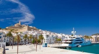 A sunny view of a port in Ibiza with a large ship docked and lots of houses and palm trees.