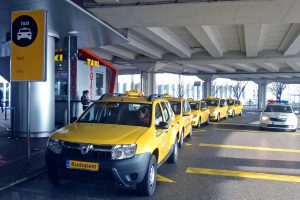 Budapest Airport taxis - Welcome Pickups A yellow Budapest Airport taxi minivan parked at the front of the taxi waiting line outside the airport exit doors.