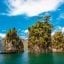 View of rock formations with trees on top in the sea at Khao Lak, surrounded by crystal waters and a blue, cloudy sky.