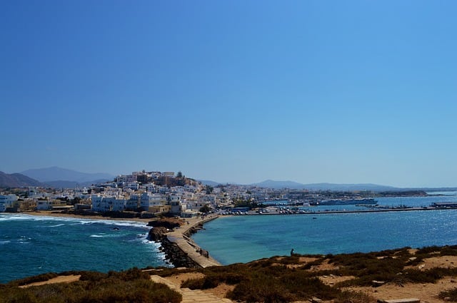 A clear daytime view of Naxos town and its coastline, with the Aegean Sea in the foreground and the island’s whitewashed buildings rising in the distance. Just a short drive from this scenic harbor lies Naxos Airport, the island’s main air gateway for domestic travelers.