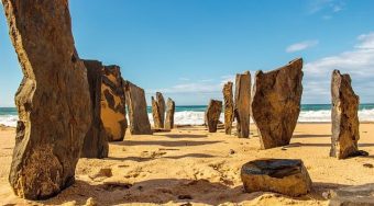 A beach in Vilamoura with large stones standing upright in the sand and blue water and skies behind.