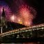 pink and red fireworks light up a dark night sky over a tall building and bridge