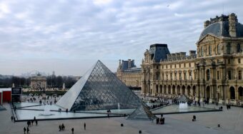 View of the Louvre Museum and glass pyramid in Paris on a cloudy afternoon, with visitors walking through the courtyard; an iconic stop during a layover in Paris.