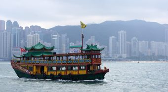 An old and colorful wooden sailboat on open water with a city in the distance.
