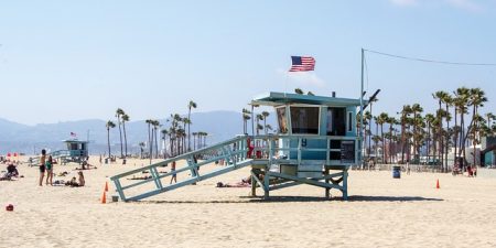 A bright image of a beach in Los Angeles, California. A light blue lifeguard booth rests in the middle of the photo with an American flag atop it. The ocean is in the background.