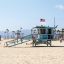 A bright image of a beach in Los Angeles, California. A light blue lifeguard booth rests in the middle of the photo with an American flag atop it. The ocean is in the background.