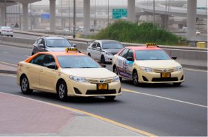 Two cream-colored Dubai taxis with orange roofs driving along a city highway in daylight, showcasing the official Dubai taxi fleet in motion amidst moderate traffic.