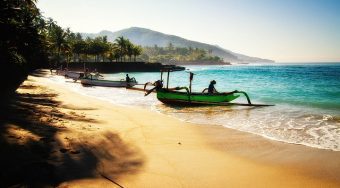 A golden sand beach in Bali lined with palm trees and local fishing boats moored onshore.