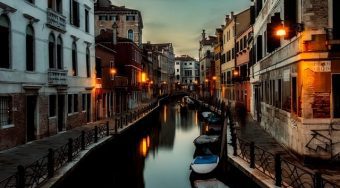 Boats docked on a canal in Venice lined with old buildings lit up at dawn.