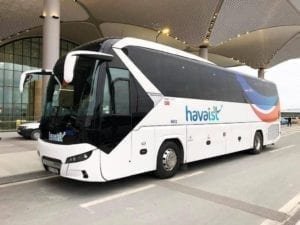 A white Havaist airport bus parked outside the airport's modern terminal building during the day, ready for passenger boarding. The sleek bus displays the “havaist” logo and provides a convenient airport transfer option.