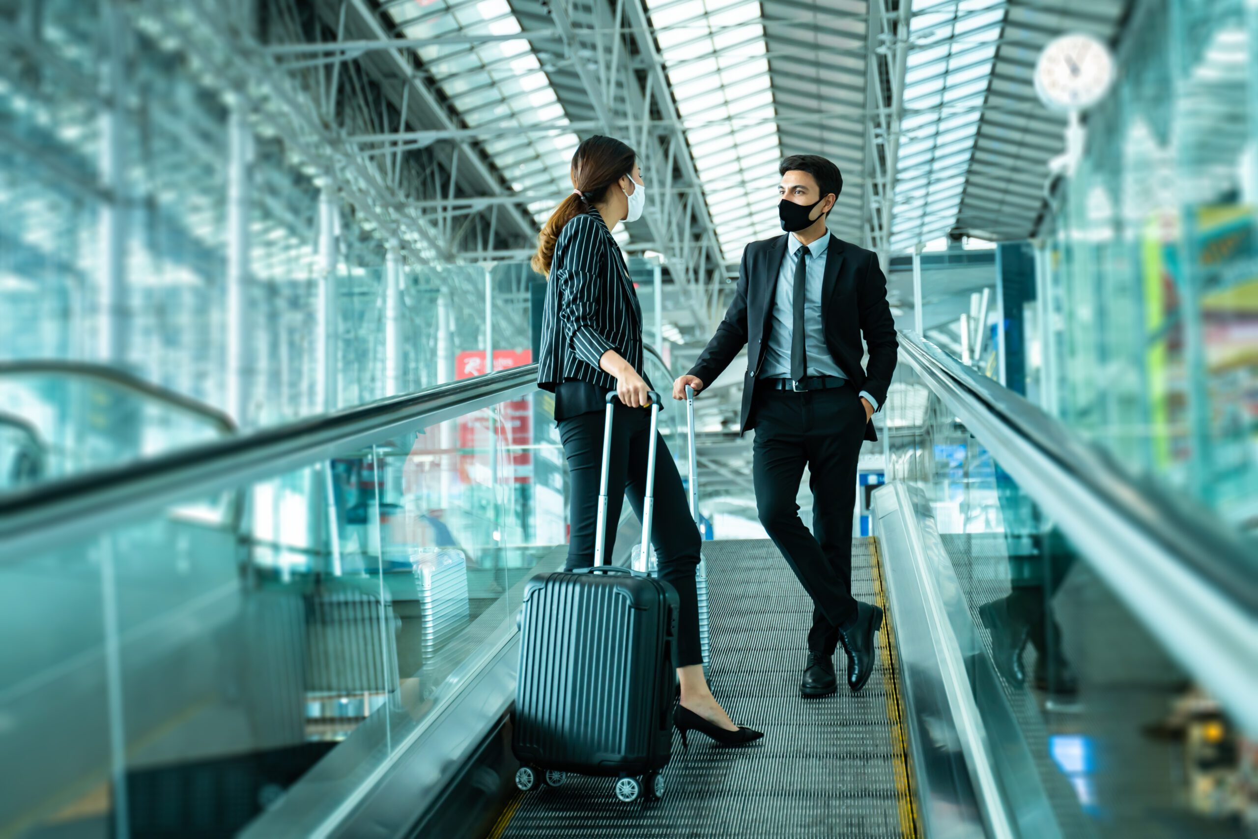 buiness man and woman wearing masks, standing on travellator in airport terminal, holding hand luggage