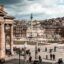 A Town square filled with people and a sandstone building with large columns.