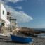 White holiday homes lining a rocky beach with wooden boats on the shore and blue skeis above.