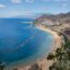 View of a large bay with a white-sand beach stretching along a mountainous coast, with blue seas and blue skies above.