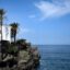 A white house with palm trees growing next to it, situated on a rocky cliff next to the sea.