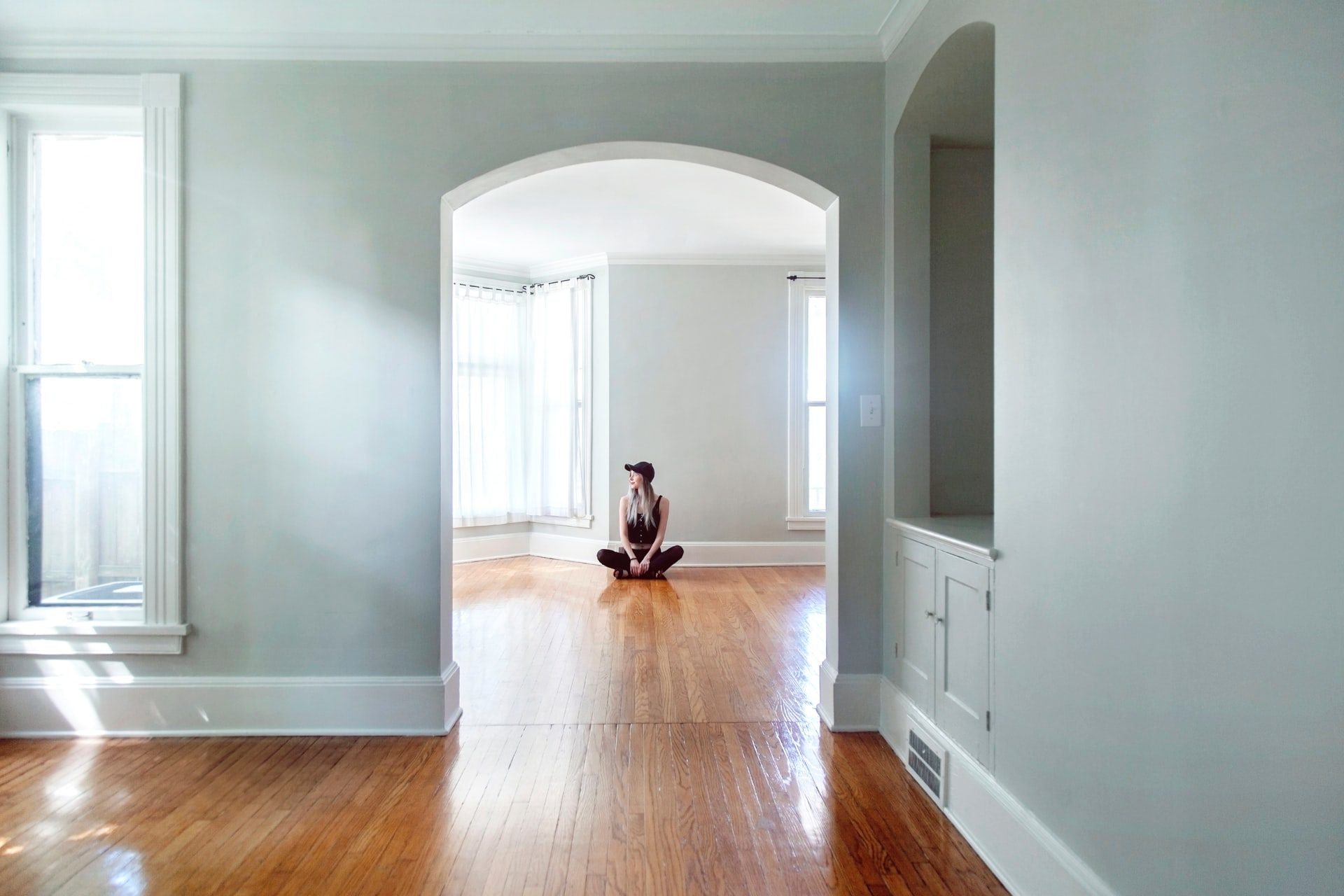 woman dressed in black and wearing baseball cap, sitting crosslegged on the floor or an empty house.