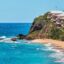blue and aqua waters crash ashore on a sandy beach with white water alongside a rocky cliff with resort homes under a blue sky