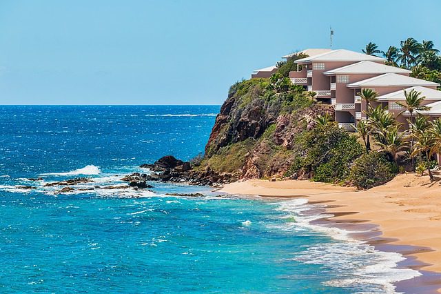 blue and aqua waters crash ashore on a sandy beach with white water alongside a rocky cliff with resort homes under a blue sky