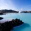 A geothermal spa with milky-blue water surrounded by black volcanic rock under a clear blue sky. Steam rises in the distance, adding to the serene and otherworldly atmosphere of the Blue Lagoon, Iceland, during daylight hours.