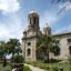 classic concrete religious building under a sunny but cloudy sky, surrounded by palm trees and green bushes