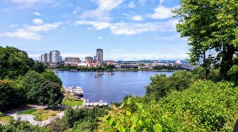 Scenic daytime view of downtown Ottawa across the river, framed by lush greenery and trees in the foreground. The skyline features modern buildings under a bright, partly cloudy sky.