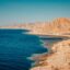 brown-beige mountains by the beach, dark blue waters under a baby blue sky