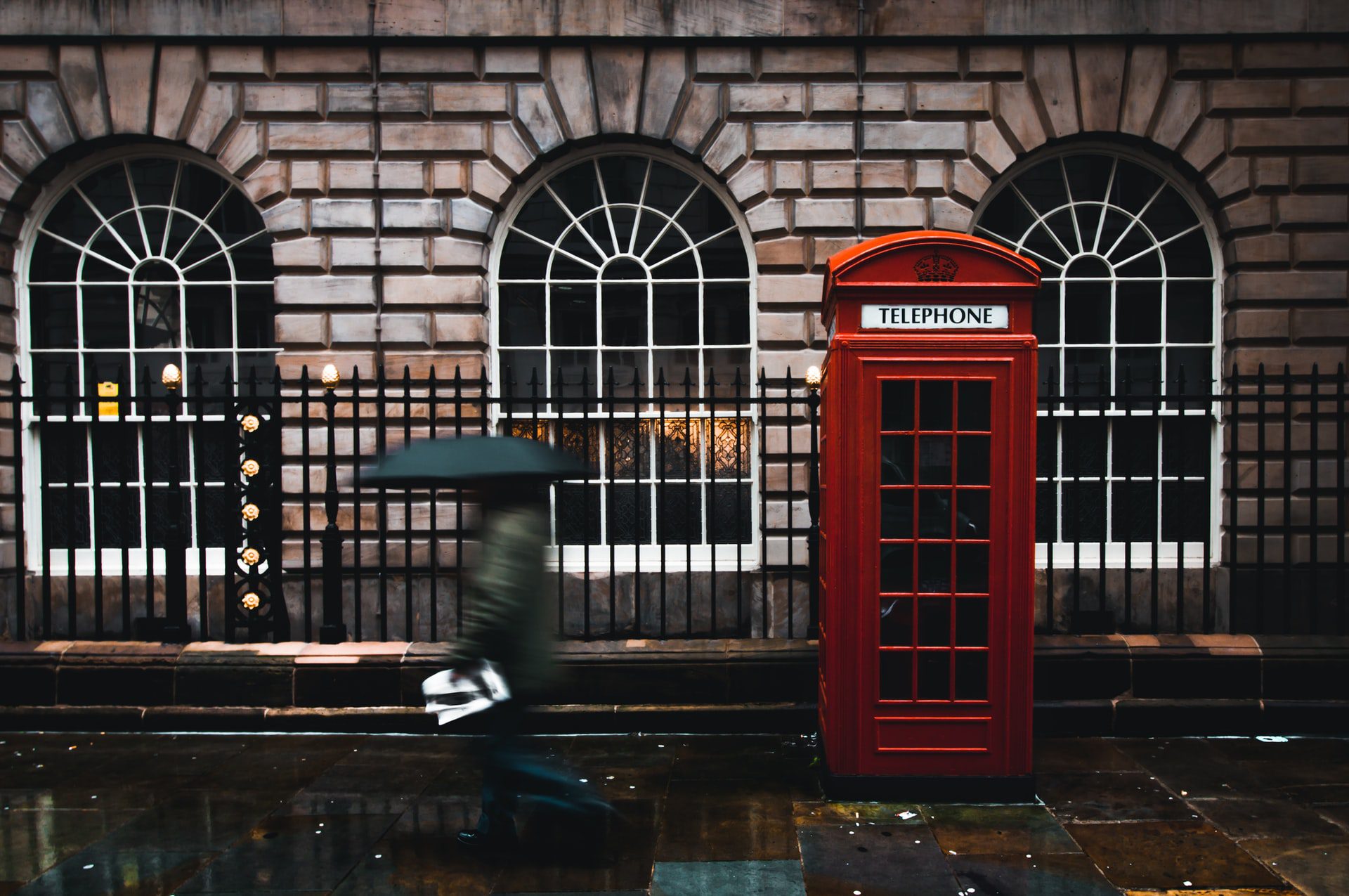 Signature red street phone booth in London with someone walking past it holding an umbrealla on a rainy day