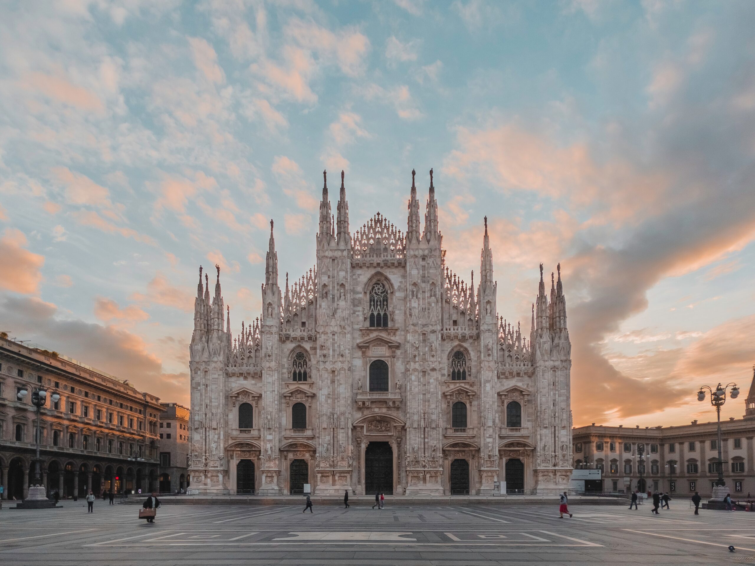 Photo of cathedral in daytime under a blue and orange sky, people walking near brown concrete building during daytime