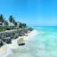 clear turquoise water by a rocky formation and some grey roof houses near palm trees in Zanzibar, Tanzania