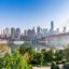 modern panoramic view of green trees by body of water, a ferry approaching a bridge and city buildings in the background under a clear blue sky during the day