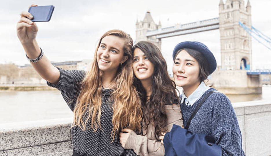 three young female travellers from different ethnic backgrounds taking a selfie with a mobile phone in front of Tower Bridge in London