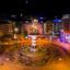 city square at nighttime, with a statue at the center, street light poles and lit up buildings surrounding it