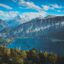 mountain range under a blue sky with some white clouds, near deep blue body of water, green trees and a green field