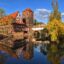 body of water shows the reflection of yellow-green trees, a bridge and a brick-red, white and brown building