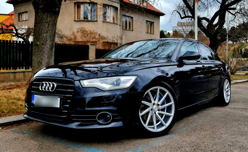 Front view of a Prague Airport transfer vehicle parked on the road outside of a house, a black Audi sedan.