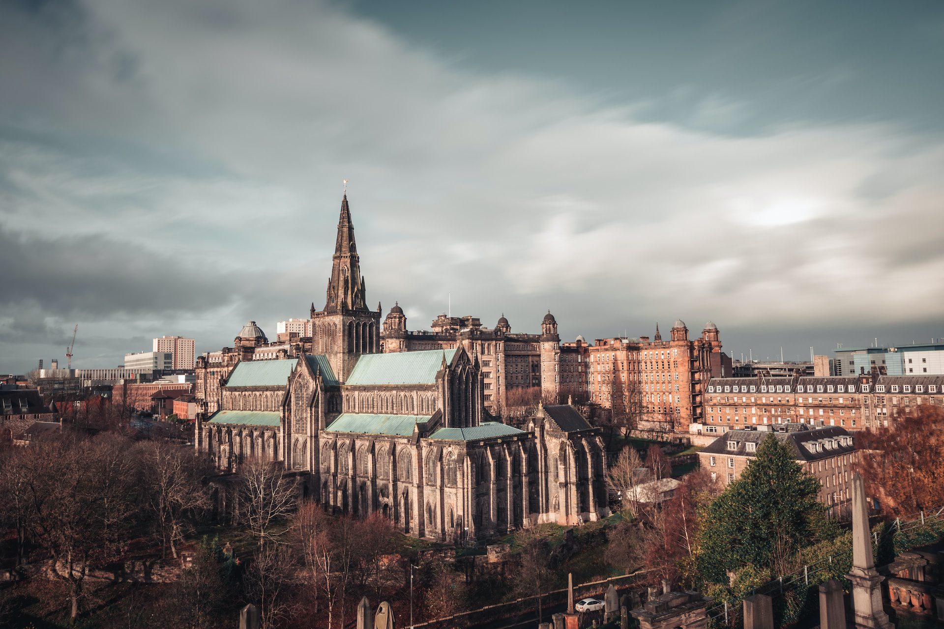 A view of Glasgow Cathedral on a cloud day, with trees to the left and city buildings behind.
