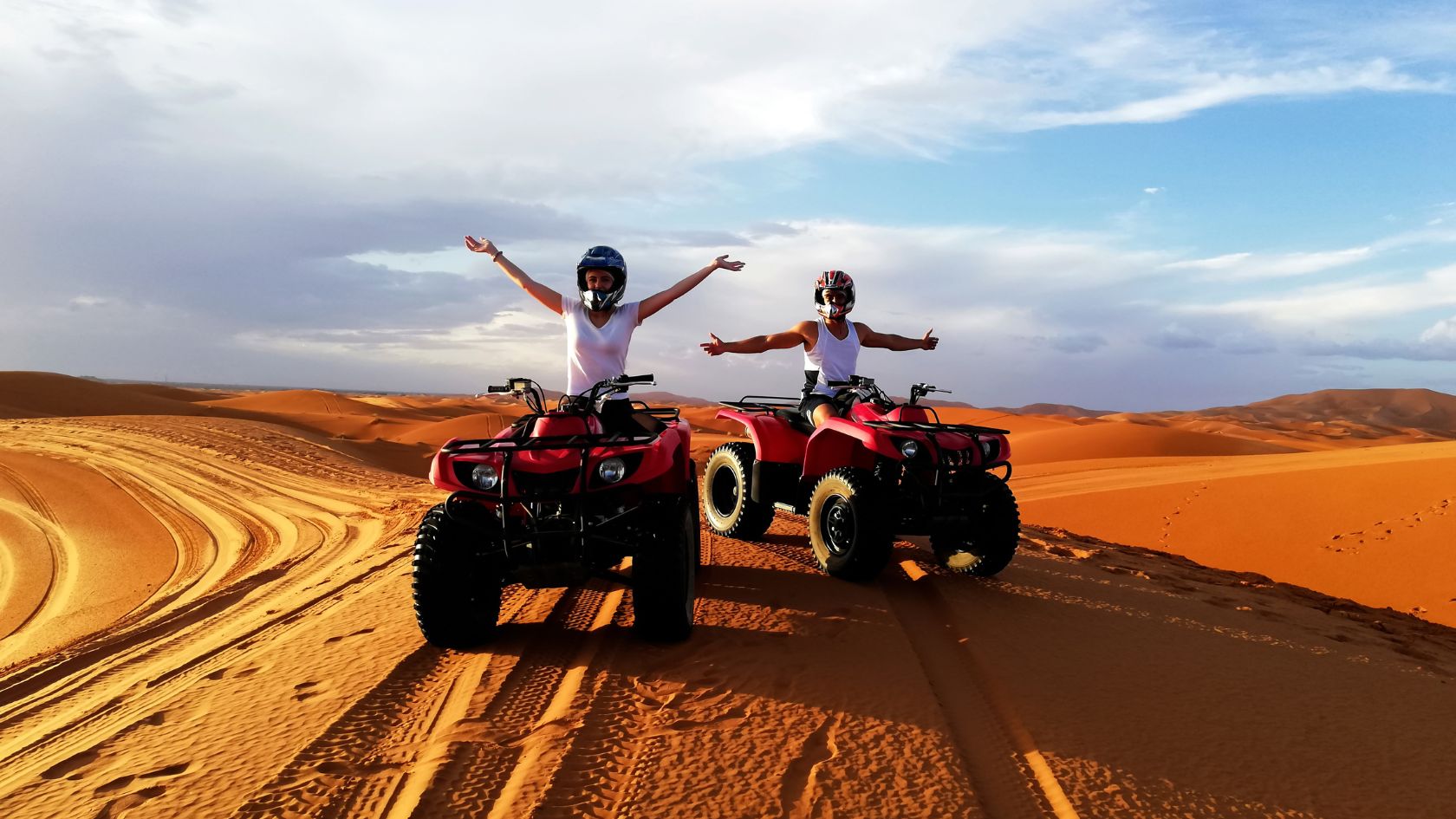 Two people on quadbikes in a desert waving their hands in the air.