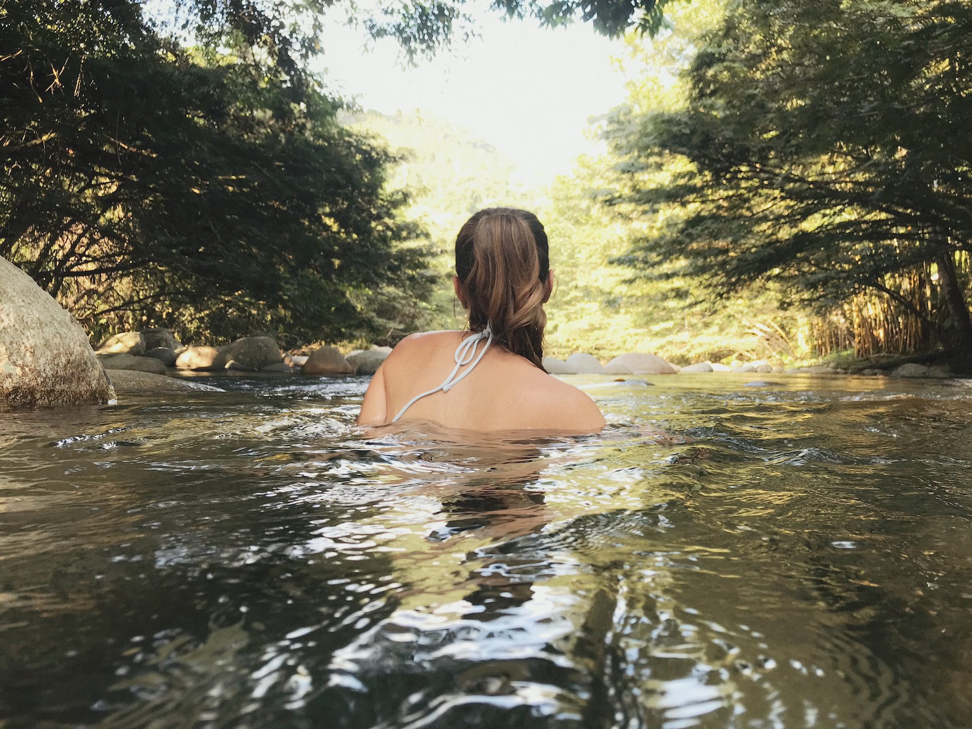 A woman submerged up to her shoulders in a swimming hole with her back to us, surrounded by trees.