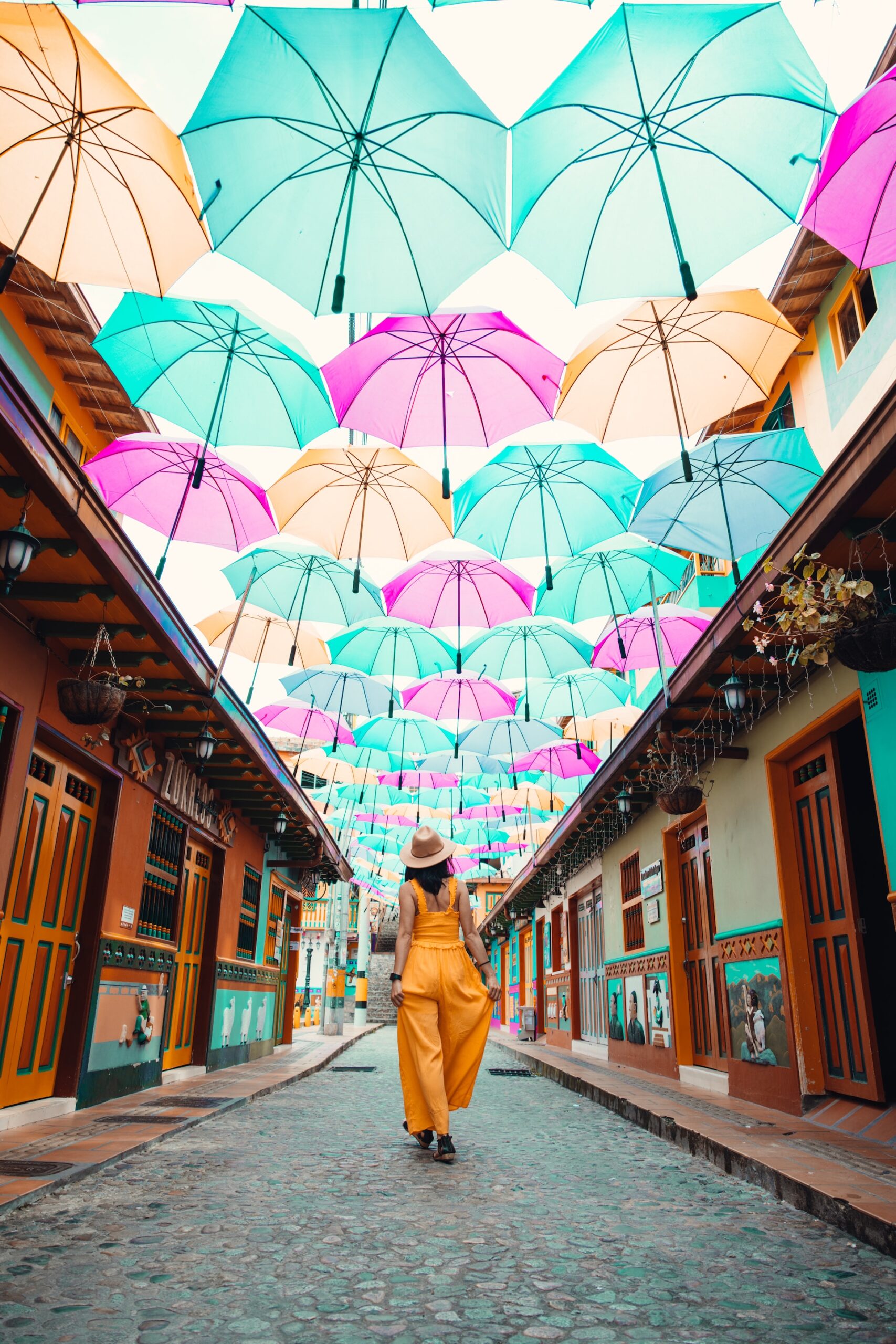 A woman walking down a cobblestoned street lined with colourful houses and with brightly coloured umbrellas hanging from above in the town of Guatape.