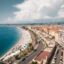 View of a palm-lined beach and the sea in Nice with a road and the city behind it and grey clouds above.