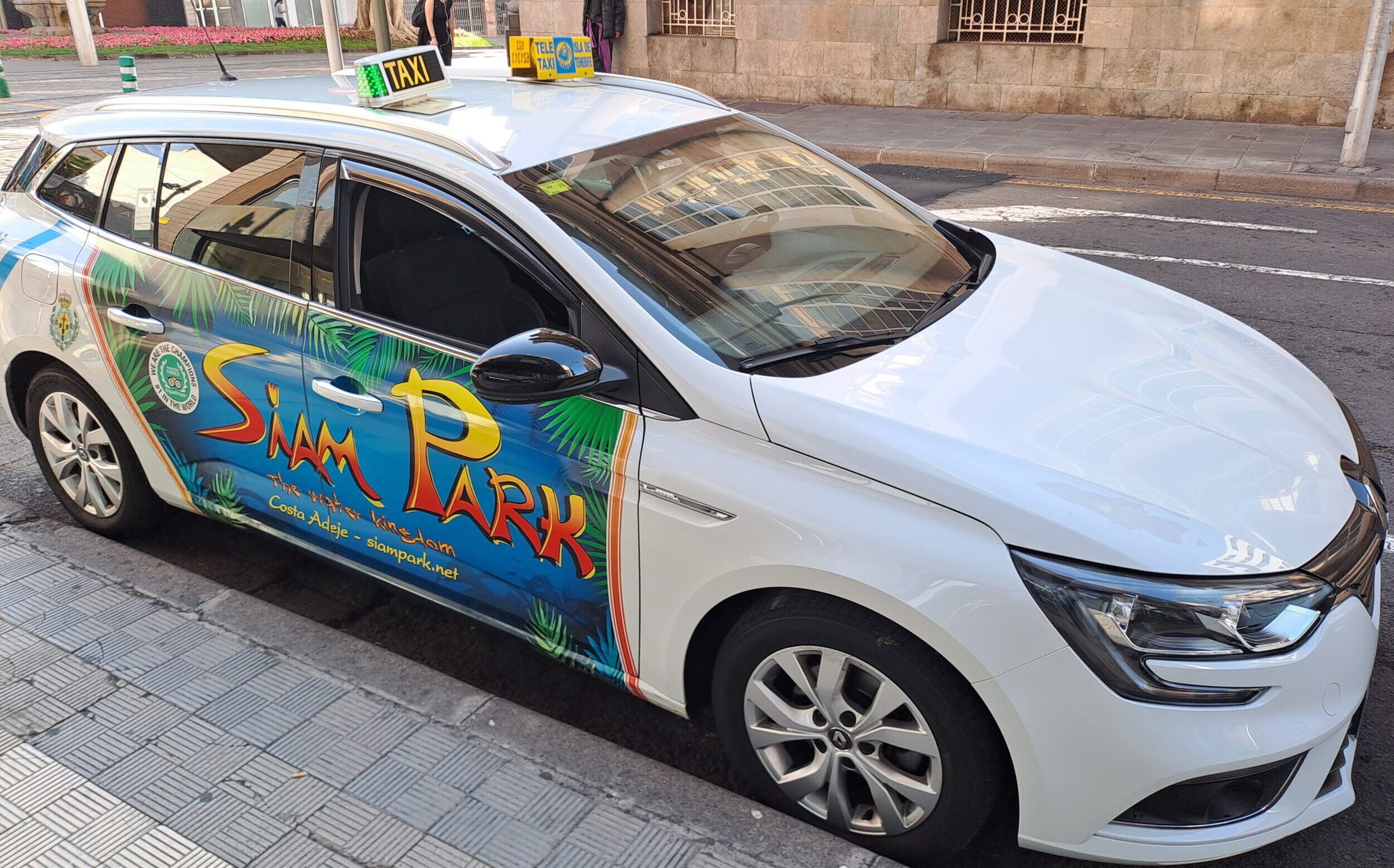 Tenerife Airport Taxi (Sedan) by Welcome Pickups Side view of a white Tenerife Airport Taxi (Sedan) with a colourful advert on the side, parked on by a curb on the street.