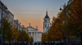 People walking down a cobbled street lined with orange-leaved trees and with Vilnius Cathedral at the end.