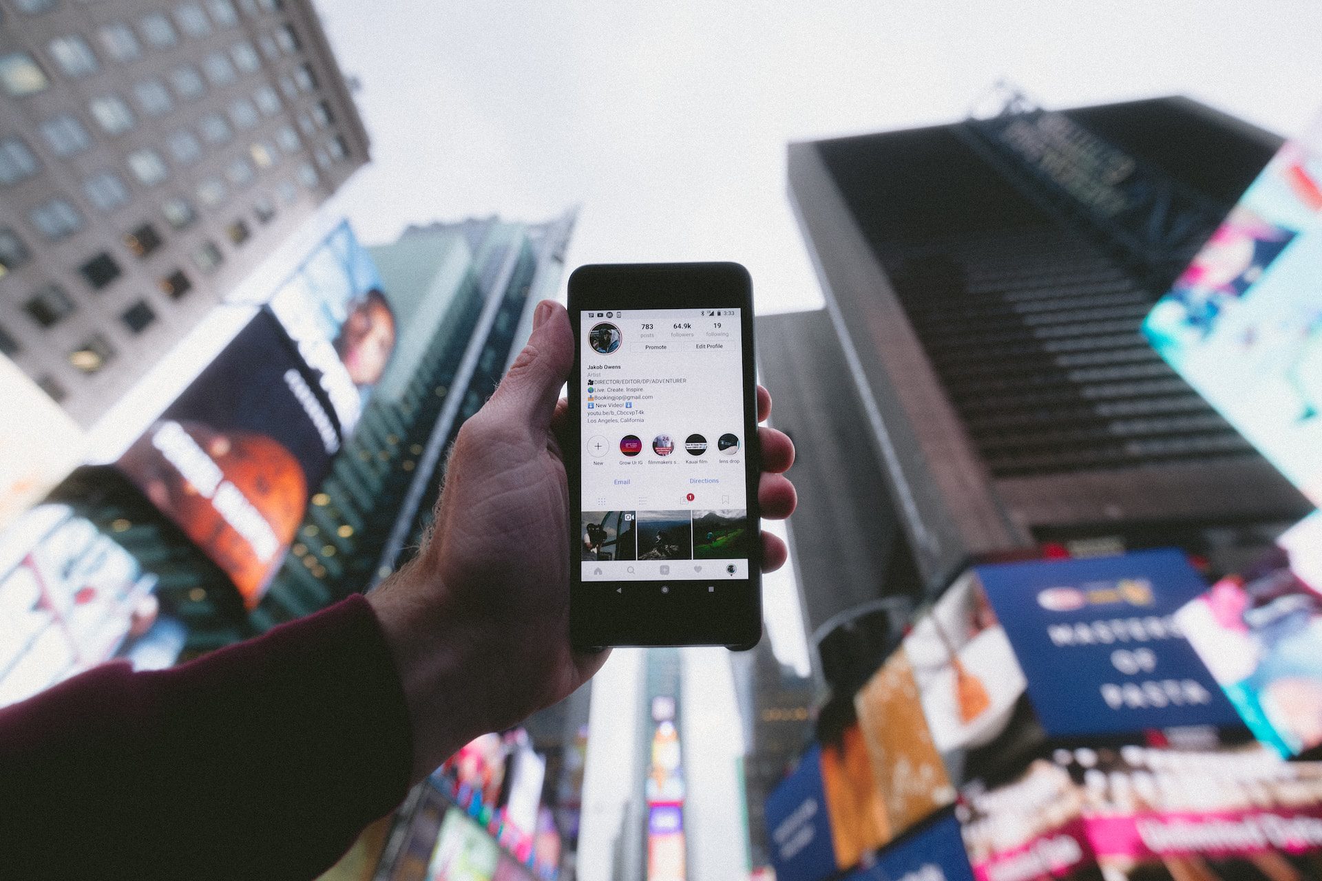 A hand holding a phone up with a social media page on the screen and skyscrapers behind.