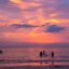 People standing in the ocean on Pantai Cenang beach at sunset, with pink and purple skies behind.
