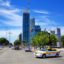 A white and yellow Montevideo taxi on a large road surrounded by trees and skyscrapers in the background.