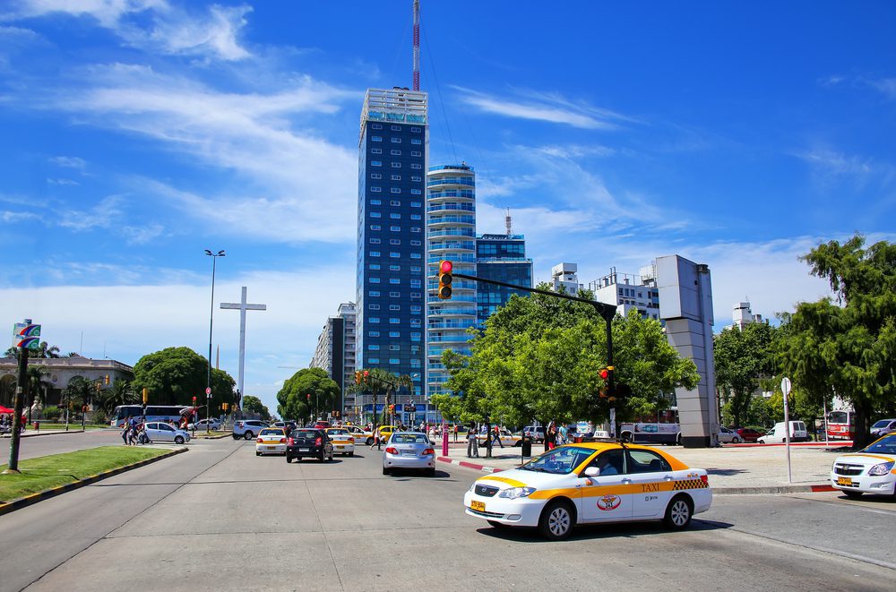Montevideo taxi A white and yellow Montevideo taxi on a large road surrounded by trees and skyscrapers in the background.