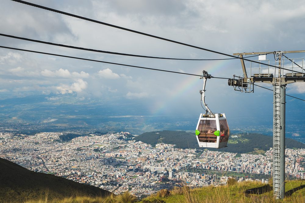Panoramic view of Quito with a cable car hanging above it and a cloudy sky above with a rainbow.