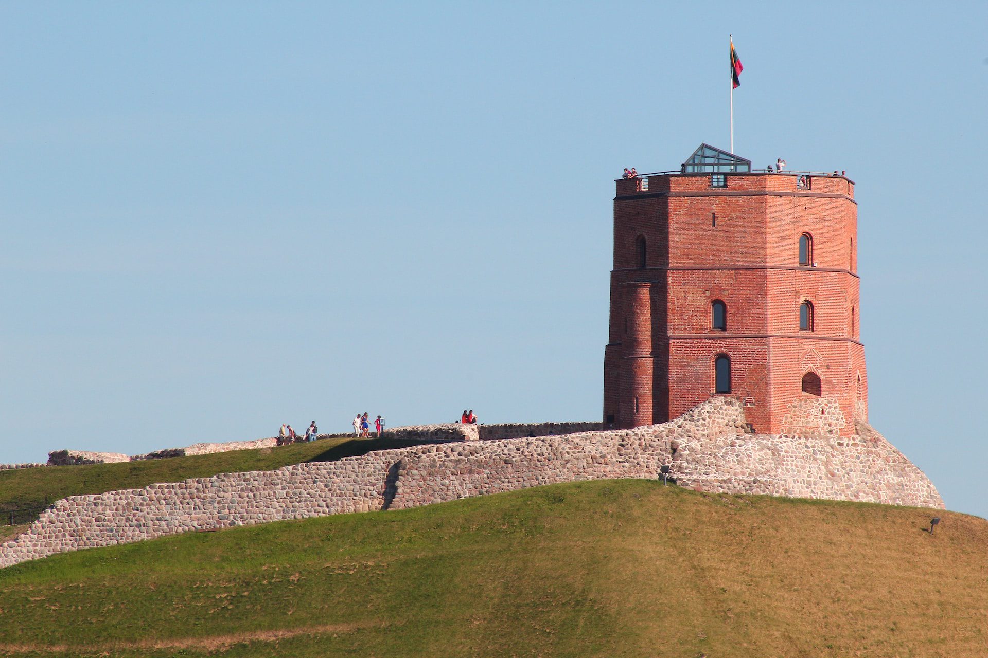 A red-brick tower standing atop a grassy hill with old castle walls around it and blue skies above.
