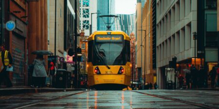 A yellow Metrolink Manchester tram on a wet street with buildings on either side.
