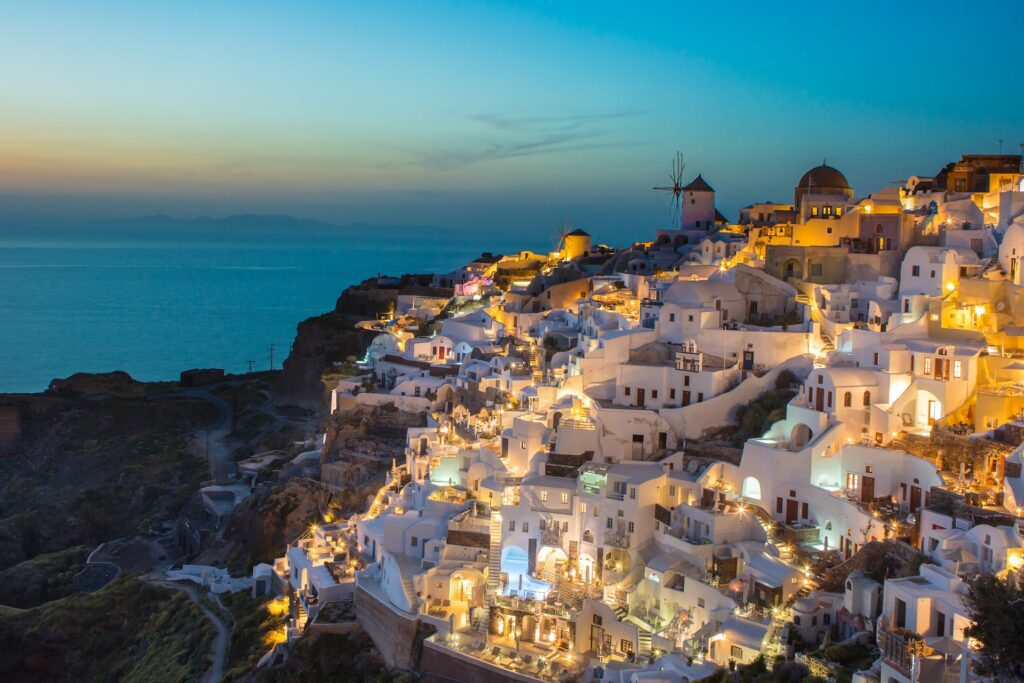 View from Oia, with lit up buildings on the cliff edge looking out towards a blue sea at dusk.
